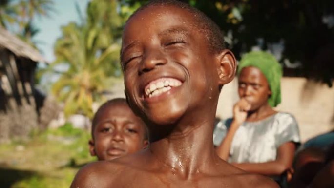 特写一个顽皮的真实的非洲孩子看着相机和其他孩子在背景中笑。快乐活力的黑人男孩与世界分享他美丽的笑容
