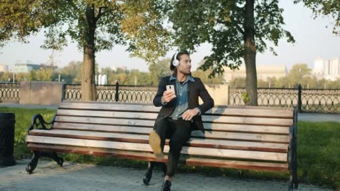 快乐的混血商人坐在城市的长凳上戴着耳机并使用智能手机的循环
