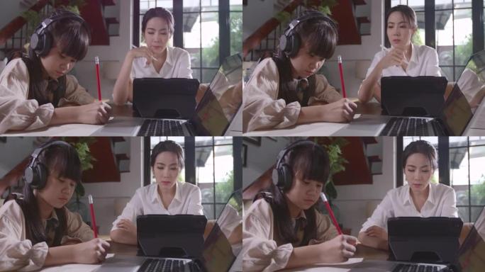 一个亚洲女孩正坐在家里专心地在线学习，在母亲的陪同下，通过平板电脑记下她学到的知识。由于Covid 