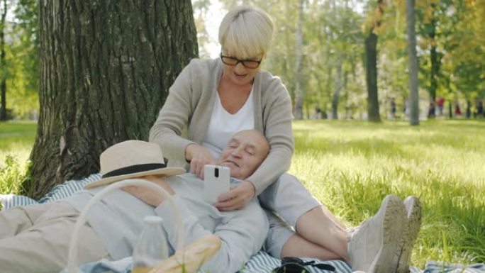 老年夫妻使用智能手机聊天在公园的草地上放松