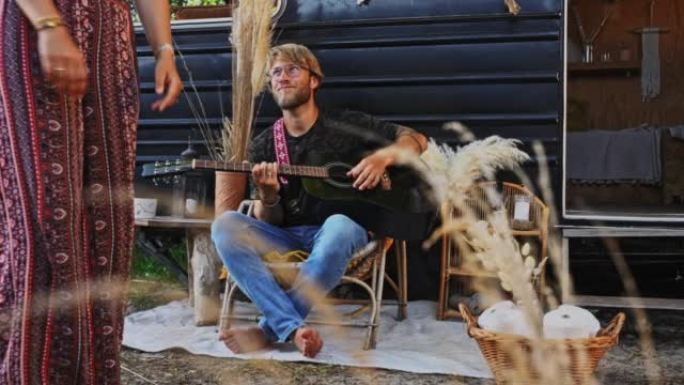 SLO MO Young hippie男人弹吉他，而女人在老式露营车前跳舞