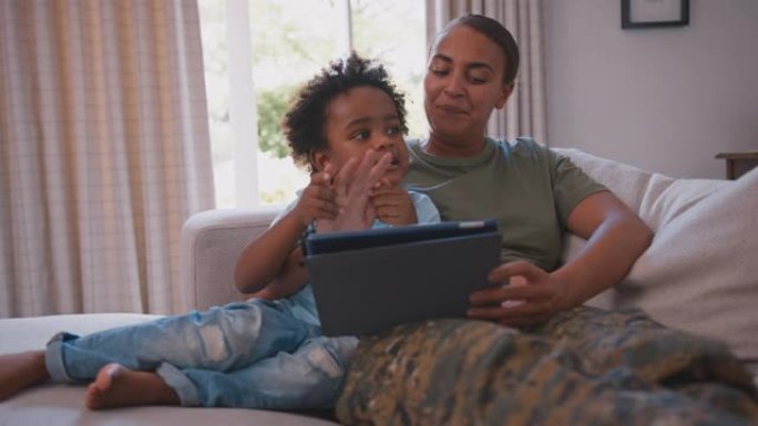 军队母亲穿着制服坐在沙发上，儿子在玩游戏或在平板电脑上播放电影——用慢动作拍摄