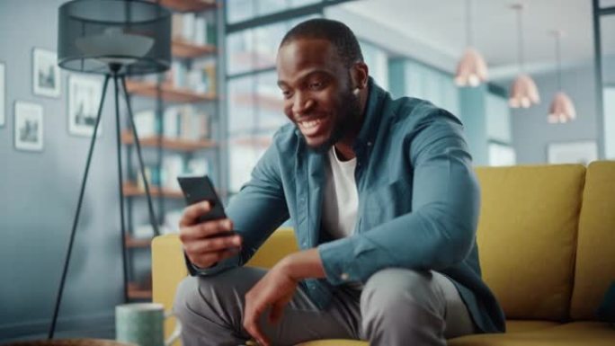 兴奋的黑人非裔美国人坐在客厅的沙发上，正在智能手机上进行视频通话。快乐的人在家里微笑，通过互联网与同
