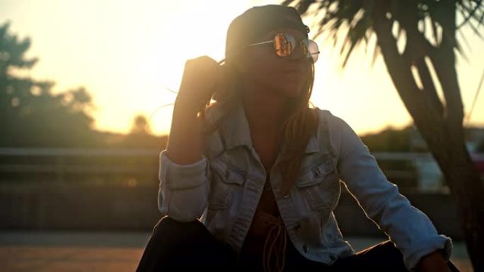 年轻女子，棕色头发，戴着帽子和牛仔夹克，在日落时分坐在木板路上的滑板上微笑，阳光
