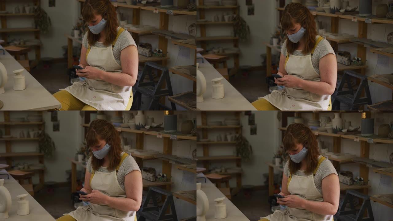 在pottery studio使用智能手机戴着口罩和围裙的女性高加索波特