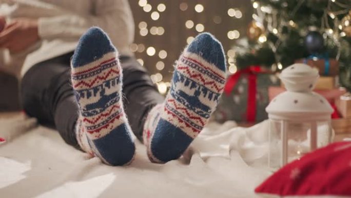 一个穿着圣诞袜的男人坐在圣诞树附近的毯子上，在手机上输入一条消息