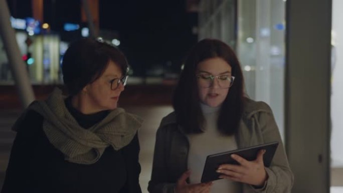 两名妇女晚上在城市使用数字平板电脑和橱窗购物