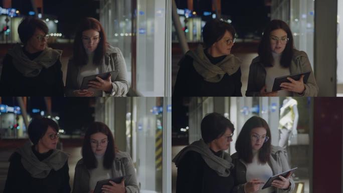 两名妇女晚上在城市使用数字平板电脑和橱窗购物