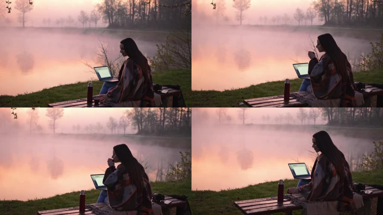 年轻女子坐在雾湖旁的长凳上喝咖啡并在笔记本电脑上工作