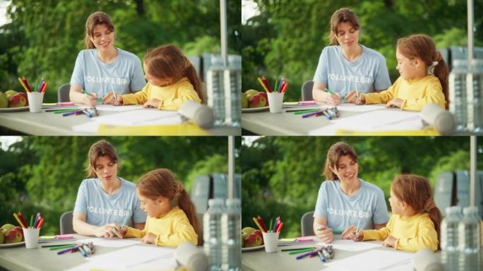 穿着黄色套头衫的才华横溢的小女孩与当地慈善组织的一名女志愿者一起画美丽的照片。人道主义援助工作者帮助