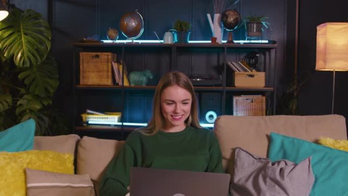 快乐美丽的金发女郎20-25自由职业女性微笑着使用笔记本电脑工作，在舒适的家中沙发上在线学习