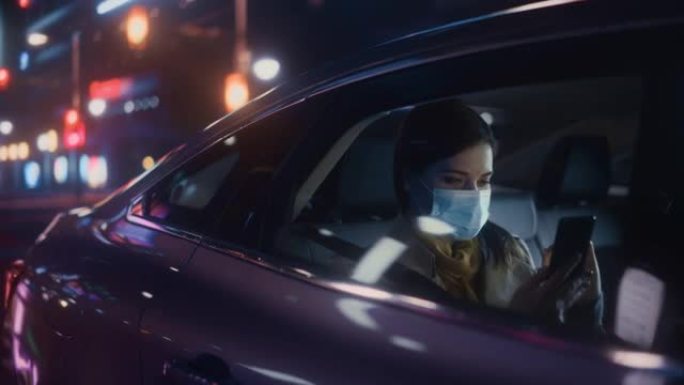 戴着口罩的女性晚上在出租车的后座上通勤回家。美丽的乘客使用智能手机，在城市街道上开着霓虹灯的汽车时看