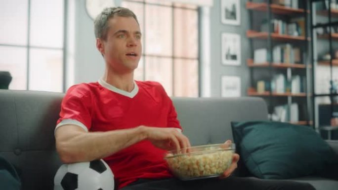 英俊的成年男子坐在沙发上观看电视足球比赛，为他的球队赢得冠军欢呼。快乐的粉丝吃零食，庆祝最喜欢的俱乐
