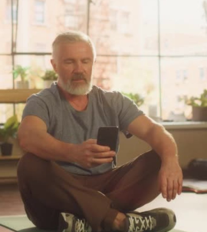 一个健康的高级男子在晨练期间在两次锻炼之间使用智能手机在公寓里的垂直特写肖像。健康的生活方式，健身，