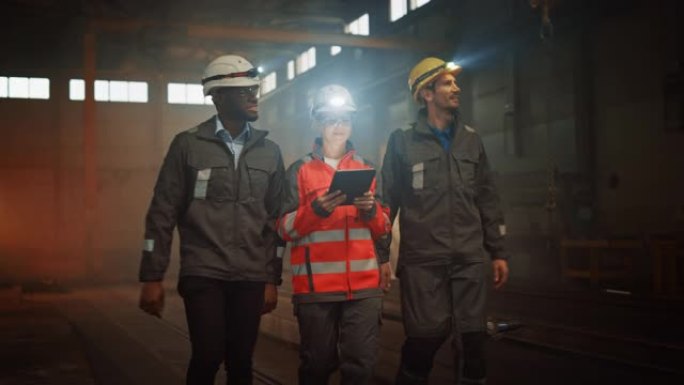 三名多元文化的重工业工程师和穿着制服的工人在黑钢工厂里用手电筒戴着安全帽行走。女性工业承包商正在使用