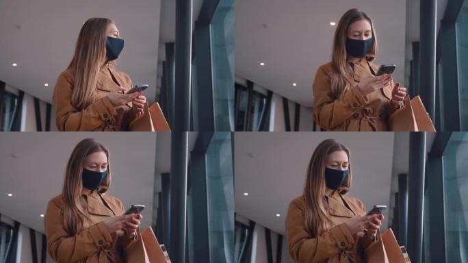 戴着口罩的快乐年轻美女在商场通道用智能手机应用程序带着购物纸袋四处张望。