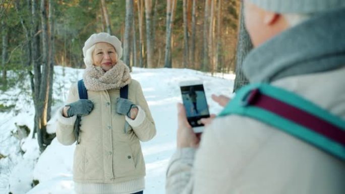 冬天，快乐的老年游客在白雪皑皑的公园为智能手机相机摆姿势