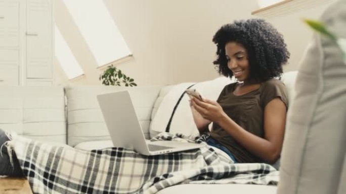 一位年轻的非洲裔美国妇女坐在家里的沙发上时使用笔记本电脑和电话。一位美丽的混血女性，非洲裔在客厅的沙