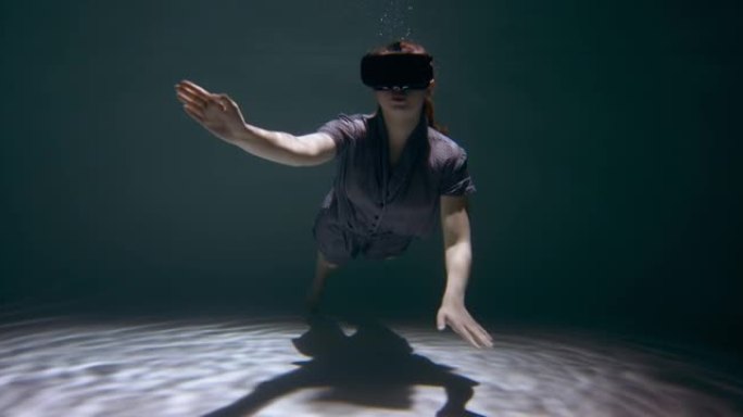 年轻漂亮的红发女人穿着睡衣，在黑暗的泳池地板上使用水下虚拟现实耳机慢动作。