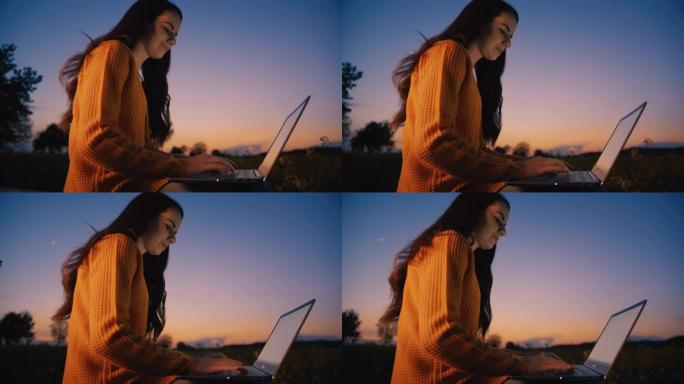 慢动作，一个中年黑发女人坐在户外在笔记本电脑上打字的特写镜头，背景是黄昏的天空