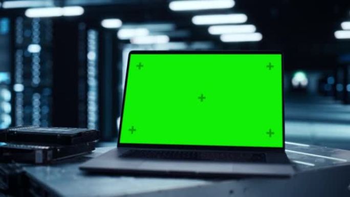 带有绿屏色度键模拟显示屏的笔记本电脑放在桌子上。在后台的现代数据中心与服务器机架的行。云计算和网络安