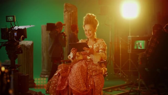 古装戏剧电影场景: 美丽的微笑女演员穿着文艺复兴时期的连衣裙，坐在椅子上，使用背景为绿屏的数字平板电