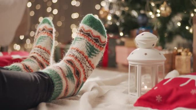 一个穿着圣诞节图案的袜子的男人坐在圣诞树附近的毯子上，用另一条腿刮擦