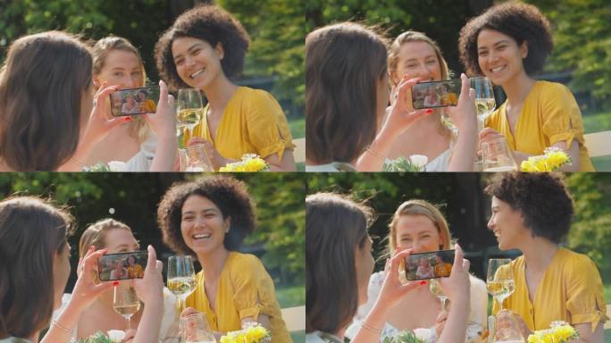 女人在坐在夏日花园里的朋友的手机上拍摄视频，一边喝着酒，一边欢呼着庆祝，一边聊天，一边追赶慢动作镜头