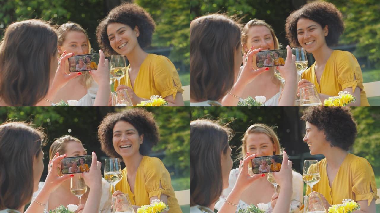 女人在坐在夏日花园里的朋友的手机上拍摄视频，一边喝着酒，一边欢呼着庆祝，一边聊天，一边追赶慢动作镜头