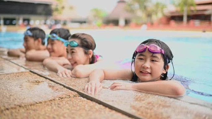 慢动作女儿看着相机微笑，而亚洲华人家庭在游泳池边休息，享受周末假期娱乐时间在游泳池里的住宿