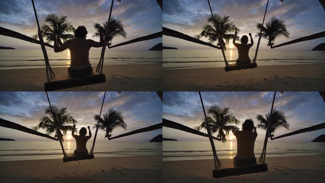 一个快乐的年轻潮人游客的剪影坐在沙滩上摇曳的秋千上，在一个热带岛屿上慢动作享受夏日日落。