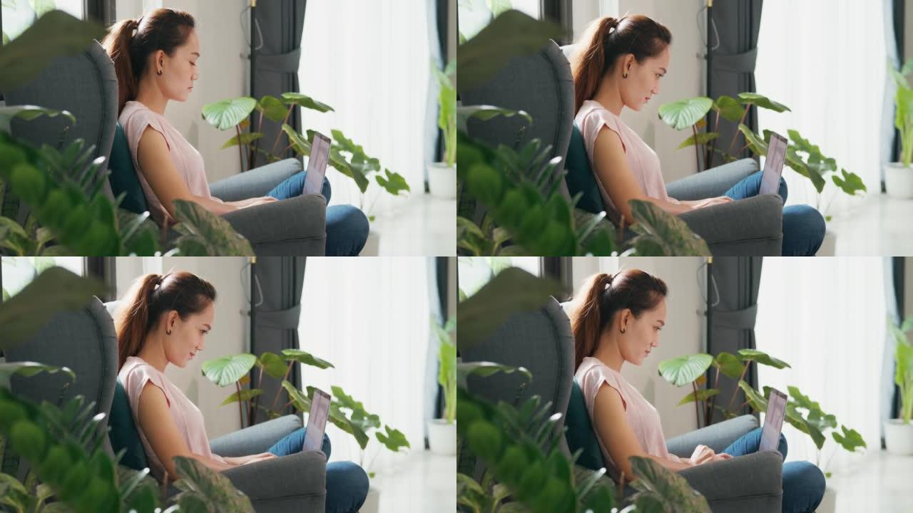 年轻的亚洲女性穿着随意平静地坐在翼椅上，使用笔记本电脑思考在客厅温室里的植物工作的想法。
