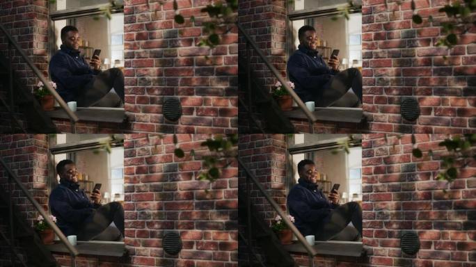 非裔美国年轻人穿着舒适的便服，使用智能手机坐在窗台上。在布鲁克林风格的褐砂石房屋中保持联系并使用互联