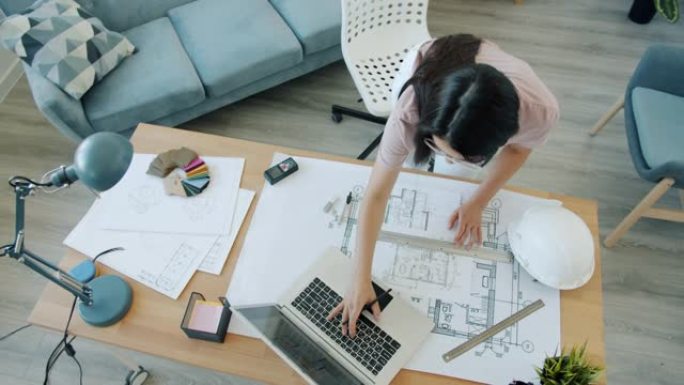 年轻的女建筑师在蓝图上绘制并在公寓室内使用笔记本电脑的高角度视图