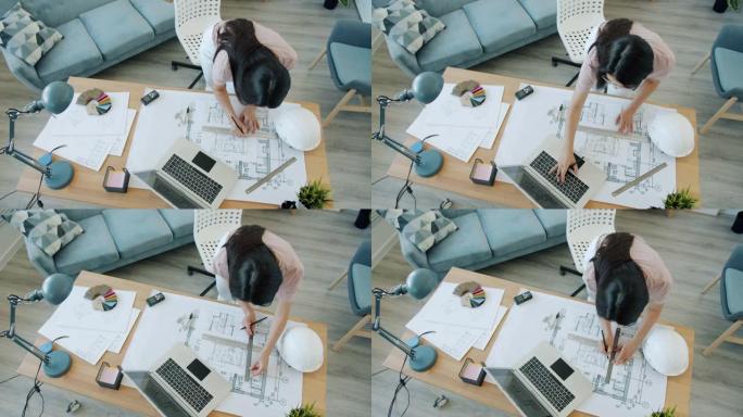 年轻的女建筑师在蓝图上绘制并在公寓室内使用笔记本电脑的高角度视图