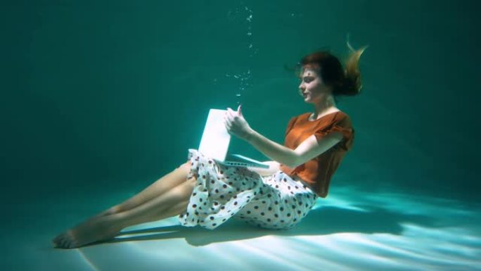 穿着休闲办公服装的美丽年轻快乐的女商人潜入水下使用笔记本电脑和慢动作。