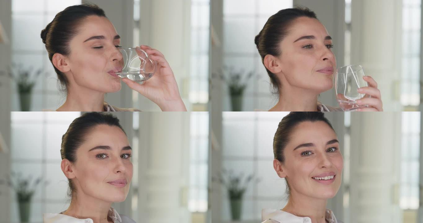 成熟微笑女人的电影镜头正在家里的客厅用透明玻璃杯喝新鲜的纯净水。她保持身体健康，感觉健康。