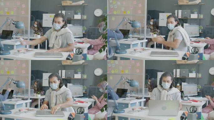 戴着口罩的员工来到工作场所问候同事消毒双手并使用笔记本电脑
