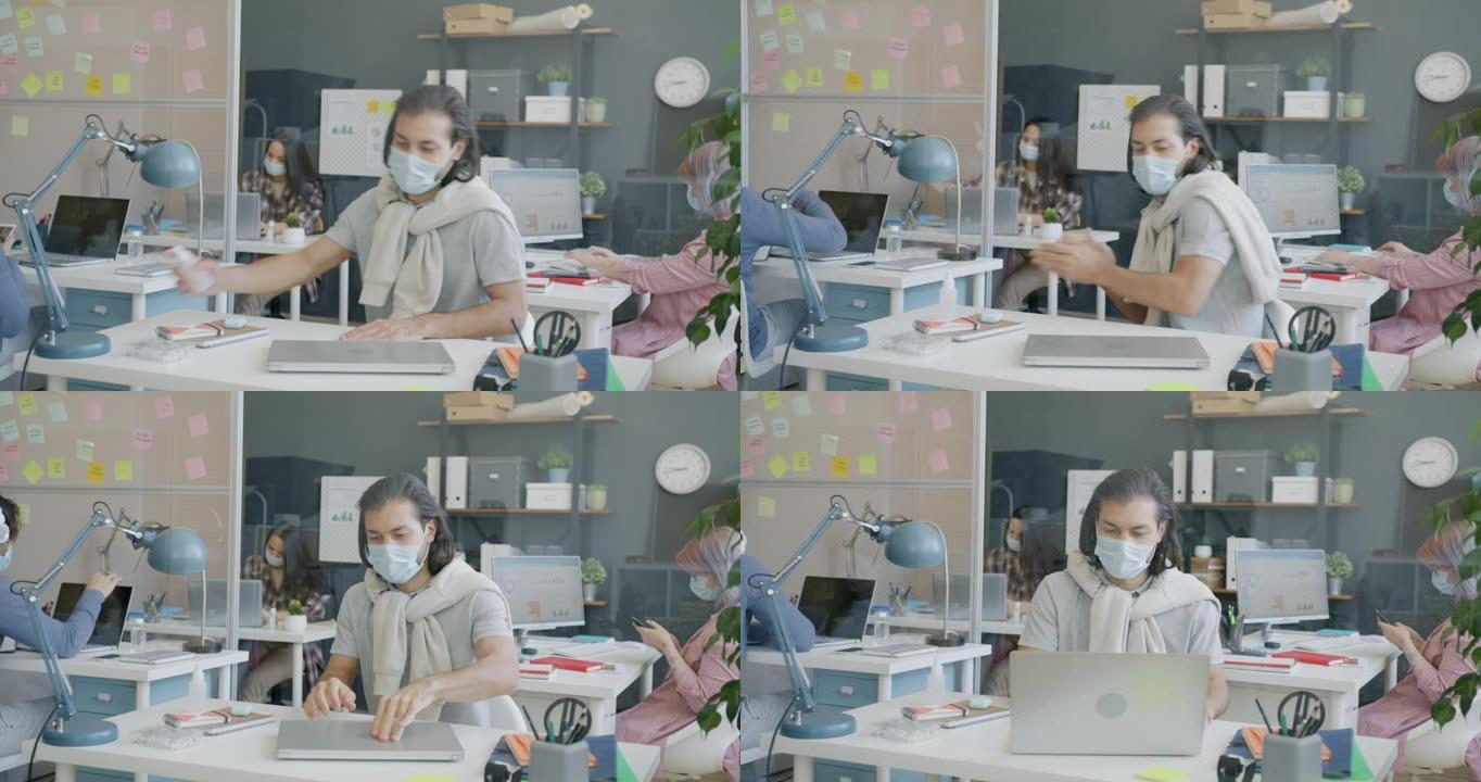 戴着口罩的员工来到工作场所问候同事消毒双手并使用笔记本电脑