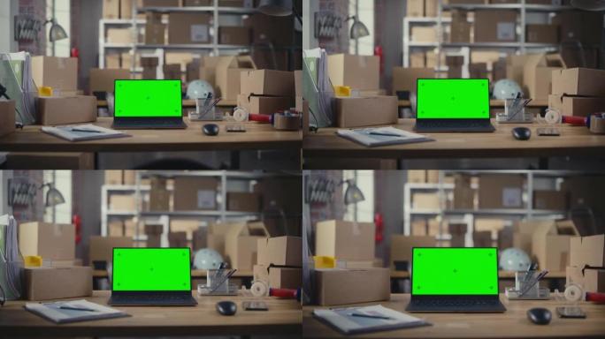 笔记本电脑站在一张绿屏Chromakey模拟显示器的桌子上。背景中有纸箱的小型企业仓库。带包裹的办公
