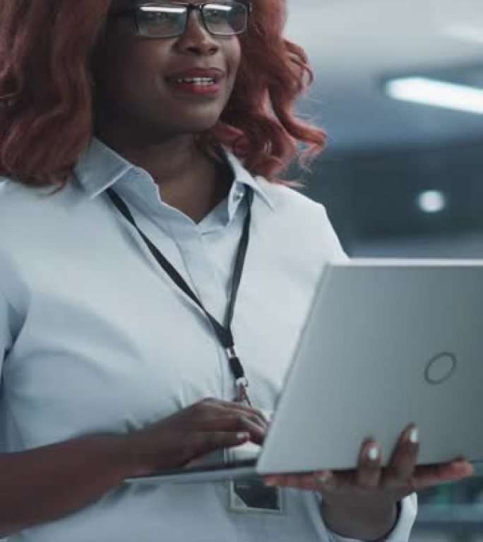 垂直屏幕: 技术软件工程部经理站立并使用笔记本电脑。黑人女性在办公室工作时看着远处思考