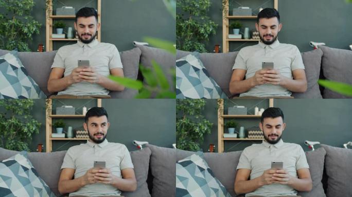 无忧无虑的阿拉伯男子在家里的沙发上使用智能手机发短信在室内浏览