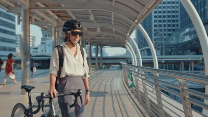 亚洲女商人戴着太阳镜去办公室上班，走路和微笑环顾四周，在城市街道上的建筑物周围拿着自行车架。