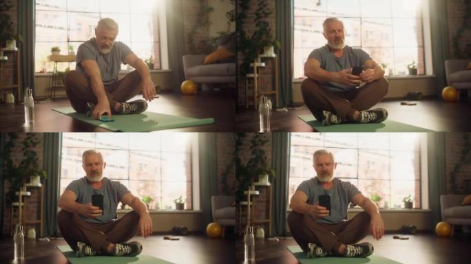 在阳光公寓的家中，在晨练期间使用智能手机进行锻炼的强壮运动健美高级男子的肖像。健康的生活方式，健身，