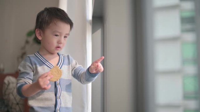 亚洲中国小男孩好奇地看着窗外，喜欢在客厅吃饼干，看着窗外