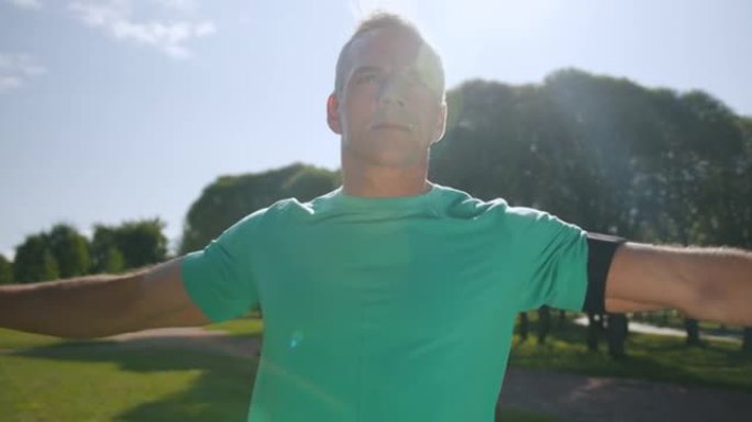 一个穿着绿松石t恤的金发健身教练正在夏天的早晨在公园里做热身运动