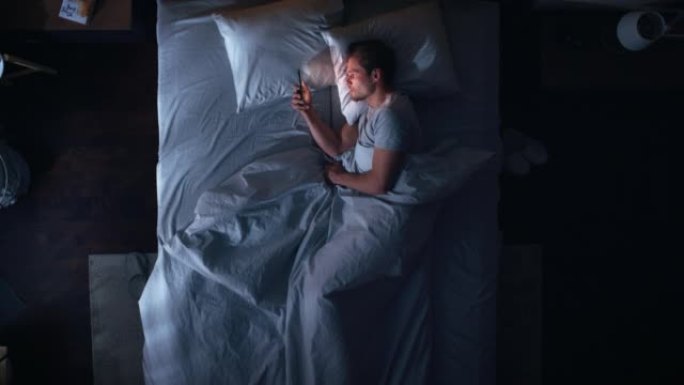 顶景公寓卧室: 英俊的年轻人晚上在床上使用智能手机。Guy浏览社交媒体，使用约会应用程序，远程工作软