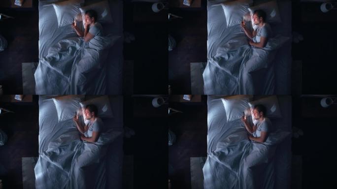 顶景公寓卧室: 英俊的年轻人晚上在床上使用智能手机。Guy浏览社交媒体，使用约会应用程序，远程工作软