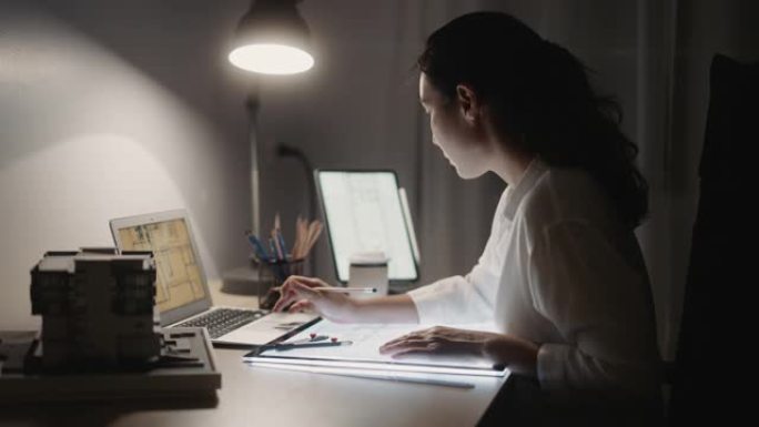 建筑师女性拿着铅笔在夜间使用笔记本电脑和蓝图进行建筑计划