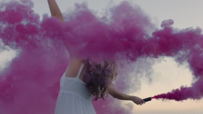 清晨，美丽的女人挥舞着粉红色的烟雾弹在海滩上跳舞，庆祝创意自由